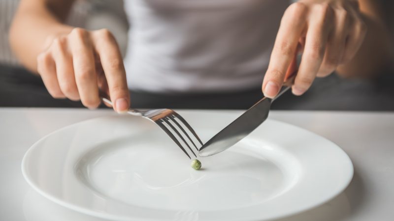 Hands hold a fork and knife over a single pea on a plate