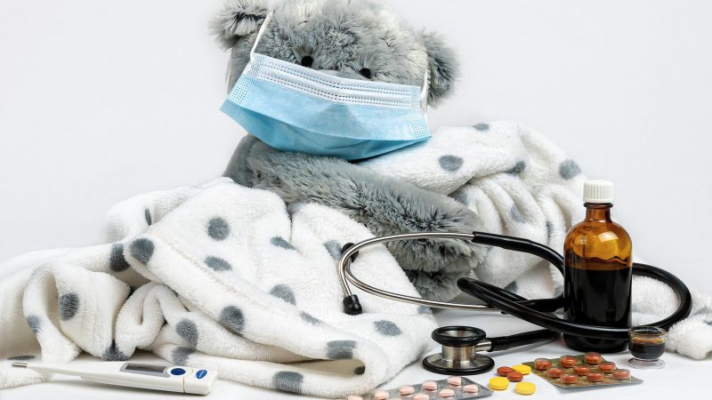 teddy bear with mask surrounded by thermometer, blanket, cough syrup, pills, and stethoscope