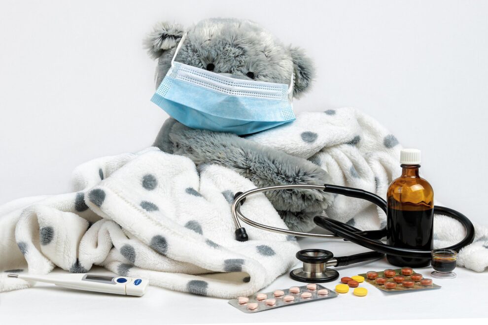 teddy bear with mask surrounded by thermometer, blanket, cough syrup, pills, and stethoscope